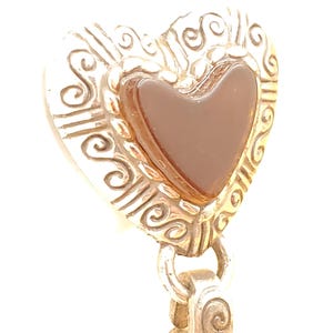 May include: A silver-coloured heart-shaped pendant featuring a brown heart-shaped stone at its centre. The pendant has an ornate, scrolled design around the edges and is attached to a decorative bail.