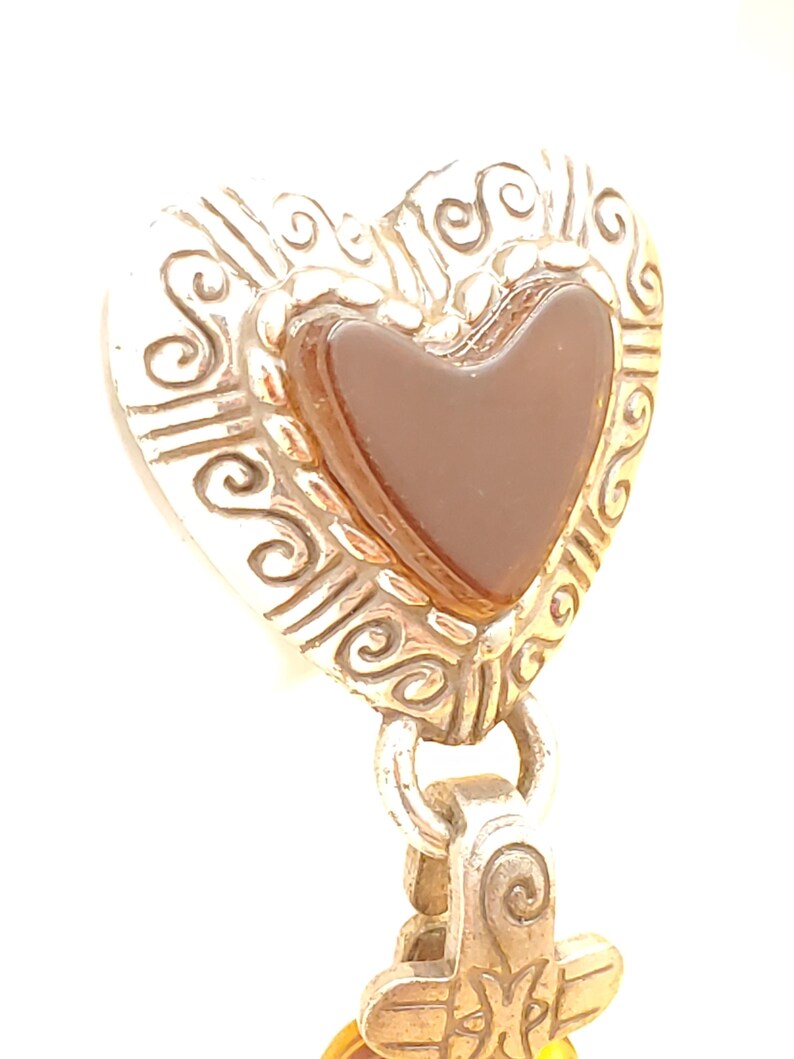 May include: A silver-coloured heart-shaped pendant featuring a brown heart-shaped stone at its centre. The pendant has an ornate, scrolled design around the edges and is attached to a decorative bail.