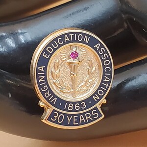 May include: A gold-colored pin with the text "VIRGINIA EDUCATION ASSOCIATION" and "30 YEARS". The pin features a central design with a torch and a small red gem. The year "1863" is also displayed.