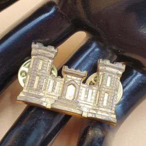 May include: A gold-coloured metal pin featuring a castle design with three towers and a central gate. The pin has two pin backs for secure attachment. The pin is likely a military or organizational emblem.