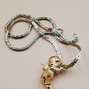 Pu&ograve; includere: Collana con pendente a forma di angelo dorato. L'angelo ha ali dettagliate e tiene in mano una pergamena. La catena &egrave; in argento e oro con un design a maglie rettangolari. La collana &egrave; esposta su uno sfondo rosa chiaro.
