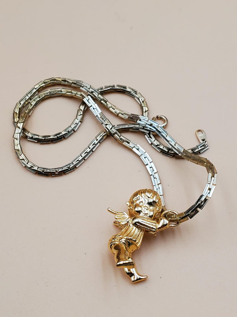 Pu&ograve; includere: Collana con pendente a forma di angelo dorato. L'angelo ha ali dettagliate e tiene in mano una pergamena. La catena &egrave; in argento e oro con un design a maglie rettangolari. La collana &egrave; esposta su uno sfondo rosa chiaro.