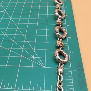 Op de afbeelding: Zilverkleurige armband met een reeks cirkelvormige schakels, elk met een textuurontwerp en goudkleurige accenten. De armband wordt getoond op een groene snijmat met inch-metingen, gelijk aan 2,54 cm.