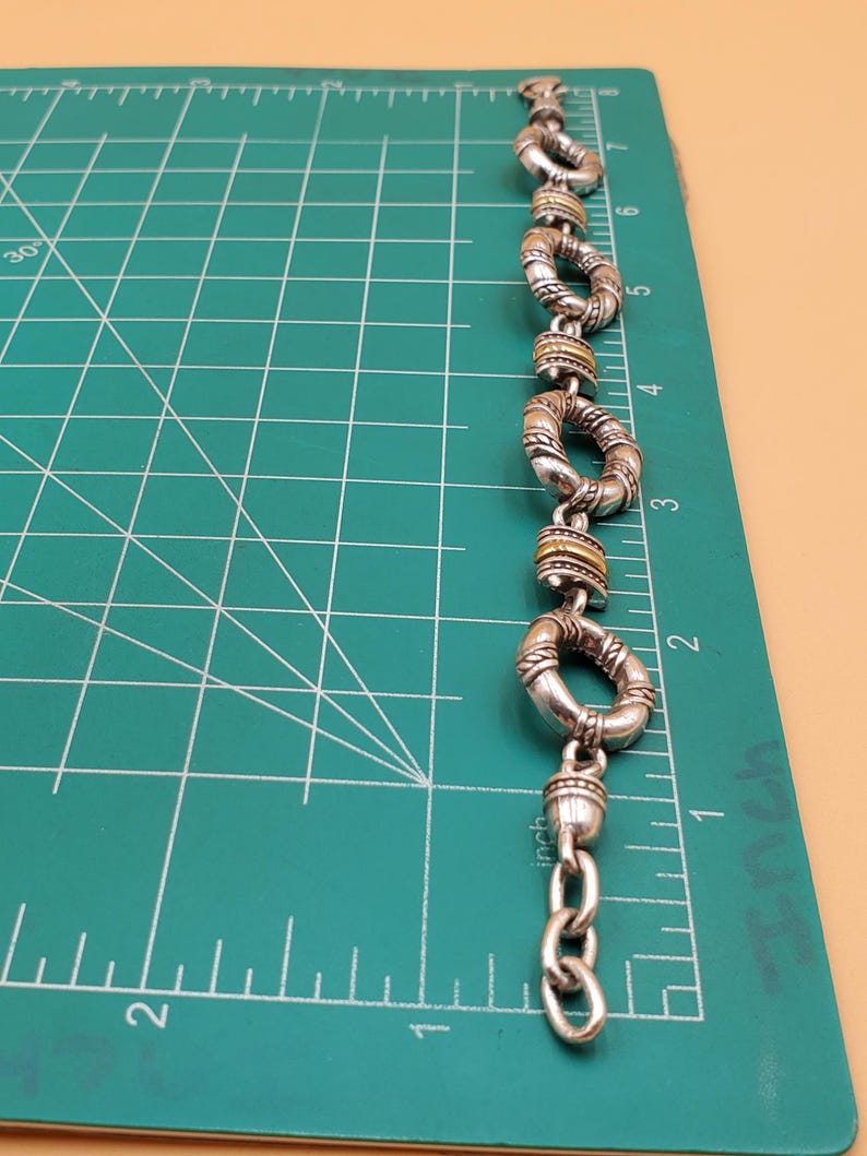 Op de afbeelding: Zilverkleurige armband met een reeks cirkelvormige schakels, elk met een textuurontwerp en goudkleurige accenten. De armband wordt getoond op een groene snijmat met inch-metingen, gelijk aan 2,54 cm.