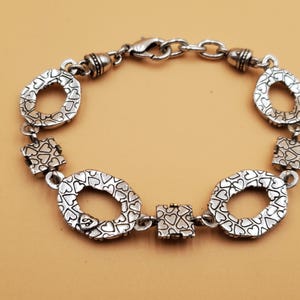 Op de afbeelding: Zilverkleurige armband met ovale en vierkante schakels. De ovale schakels hebben een hartvormig patroon en de vierkante schakels zijn bedekt met hartjes. Met een karabijnsluiting.