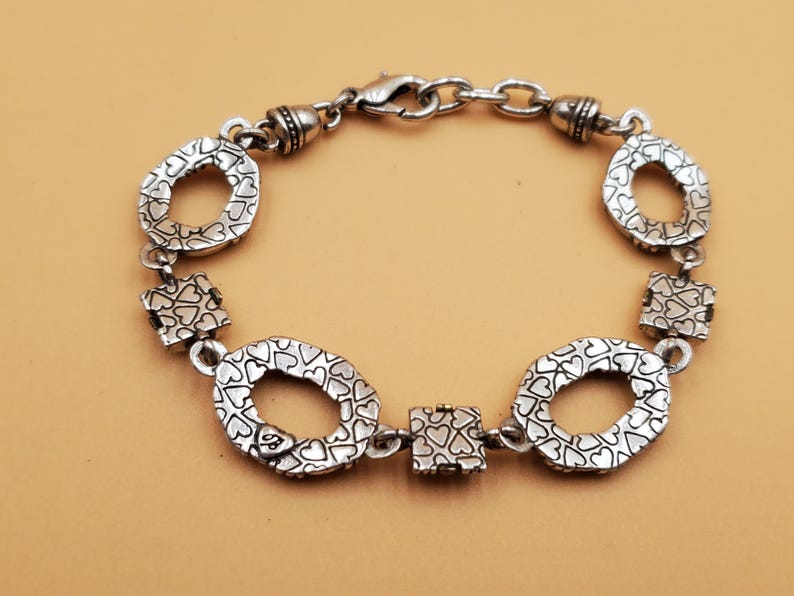 Op de afbeelding: Zilverkleurige armband met ovale en vierkante schakels. De ovale schakels hebben een hartvormig patroon en de vierkante schakels zijn bedekt met hartjes. Met een karabijnsluiting.