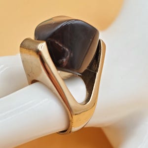 Puede incluir: Un anillo dorado con una gran gema facetada de color marrón oscuro. El anillo tiene un diseño único y abierto con una banda ancha y soportes curvos para la piedra. La gema es un punto focal prominente.