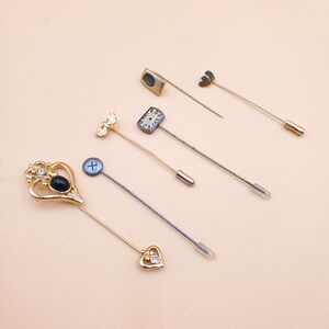 May include: An assortment of decorative lapel pins in various designs. The pins feature gold and silver tones, with embellishments including a heart, a butterfly, a cross, and a small clock face. Some pins have clear plastic tips.