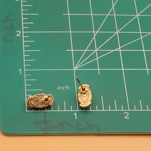 May include: Gold-toned stud earrings with an oval design and intricate detailing. The earrings are approximately 0.75 inches long and are displayed on a green cutting mat with inch measurements.