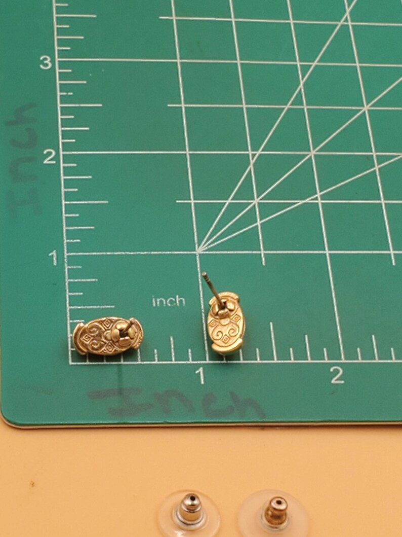 May include: Gold-toned stud earrings with an oval design and intricate detailing. The earrings are approximately 0.75 inches long and are displayed on a green cutting mat with inch measurements.