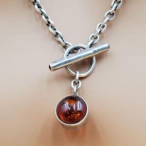 May include: A silver necklace with a toggle clasp and a pendant featuring a round, amber-colored stone. The chain is made of interlocking links. The pendant is a silver bezel setting around the amber stone.