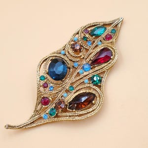May include: A gold-toned brooch in the shape of a leaf, adorned with colorful rhinestones. The brooch features a variety of colors, including blue, green, red, and brown.