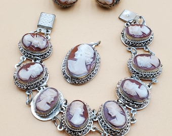 May include: A jewelry set featuring a cameo design. The set includes a bracelet, pendant, and two rings. Each piece has a silver-colored metal frame with a detailed cameo portrait in shades of brown and white.