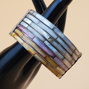 May include: A silver-toned bracelet with a rectangular design. The bracelet is inlaid with rectangular pieces of mother-of-pearl in shades of white, pink, yellow, and iridescent hues. The bracelet is displayed on a black hand.