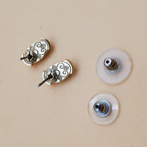 May include: Close-up of two gold-colored stud earrings with decorative patterns and two clear earring backs. The earrings have a post and butterfly backing. The background is a light tan color.
