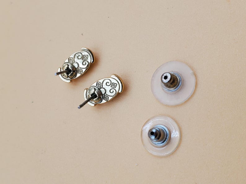 May include: Close-up of two gold-colored stud earrings with decorative patterns and two clear earring backs. The earrings have a post and butterfly backing. The background is a light tan color.
