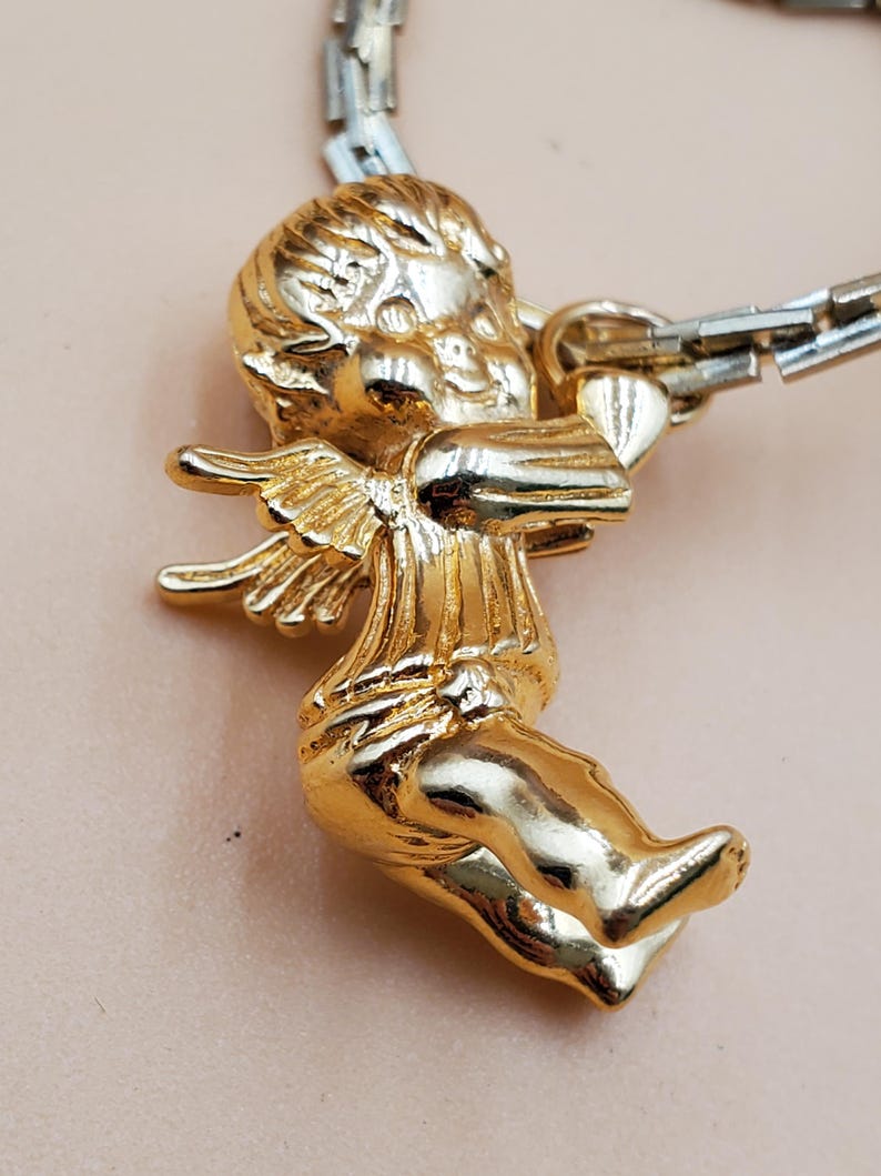 Pu&ograve; includere: Un ciondolo a forma di cherubino dorato con ali spiegate e un arco. Il cherubino &egrave; attaccato a una catena color argento. Il ciondolo &egrave; molto dettagliato e ha una finitura lucida.