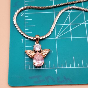May include: A gold-coloured necklace with an angel pendant. The pendant has a pear-shaped pink gemstone, a round clear gemstone, and gold-coloured wings. The necklace is on a green surface with inch measurements.