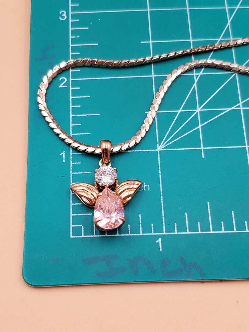 May include: A gold-coloured necklace with an angel pendant. The pendant has a pear-shaped pink gemstone, a round clear gemstone, and gold-coloured wings. The necklace is on a green surface with inch measurements.