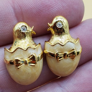 May include: A pair of gold tone earrings shaped like baby chicks hatching from eggs. The eggs are white with a gold trim and a gold bow. Each chick has a rhinestone eye.