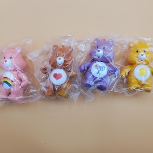 May include: Four Care Bear figurines in plastic packaging. The bears are pink, brown, purple, and yellow. Each bear has a unique belly badge: a rainbow, a heart, a lollipop, and a sun. The toys are on a light brown surface.