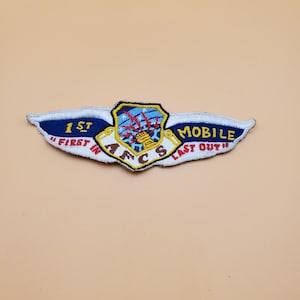 May include: Embroidered patch with a shield and wing design. The patch features the text "1st First In" on the left wing and "Mobile Last Out" on the right wing. The shield has a blue background with red lightning bolts and a gold structure.