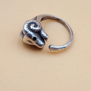 May include: A silver-toned ring featuring a ram's head with detailed horns and a textured surface. The ring has an open design, allowing for adjustable sizing. The ram's head is the focal point, with a dark eye detail.