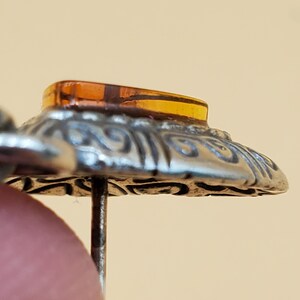 May include: Close-up of a vintage hat pin. The pin features a silver-toned, ornate, circular base with an amber-coloured, rectangular stone at the top. The pin is held by a finger.
