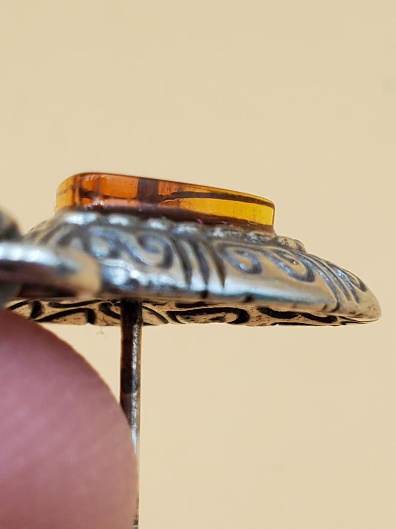 May include: Close-up of a vintage hat pin. The pin features a silver-toned, ornate, circular base with an amber-coloured, rectangular stone at the top. The pin is held by a finger.