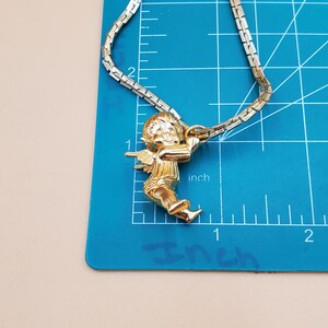Pu&ograve; includere: Collana dorata con un ciondolo a forma di cherubino che tiene un arco. La catena &egrave; a maglie piatte. Il ciondolo &egrave; alto circa 2,5 cm. La collana &egrave; appoggiata su un tappetino da taglio blu con misure in pollici.