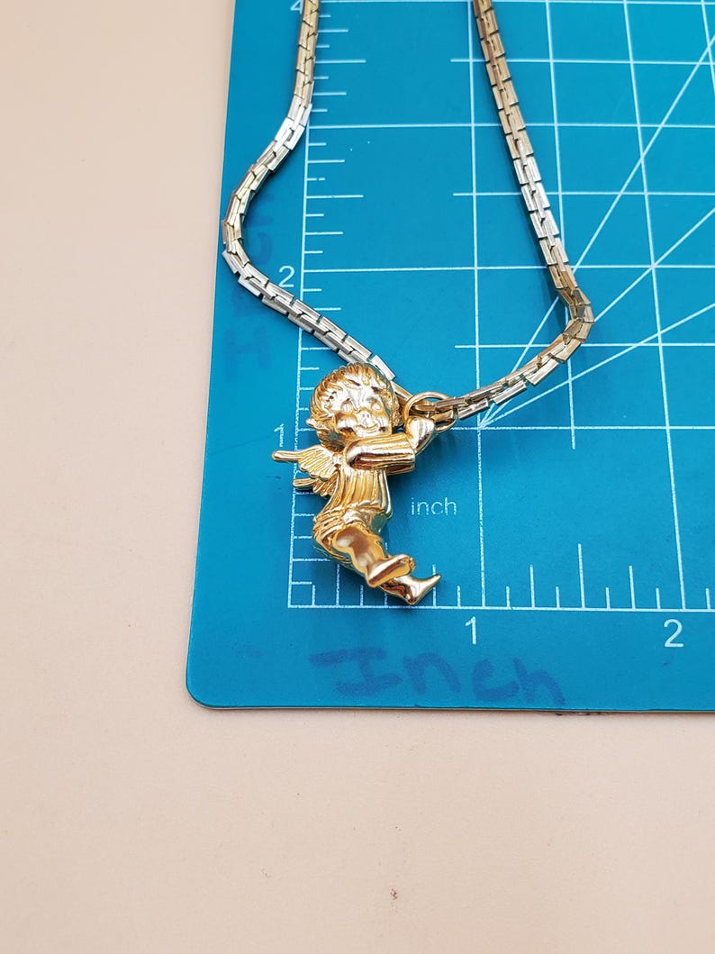 Pu&ograve; includere: Collana dorata con un ciondolo a forma di cherubino che tiene un arco. La catena &egrave; a maglie piatte. Il ciondolo &egrave; alto circa 2,5 cm. La collana &egrave; appoggiata su un tappetino da taglio blu con misure in pollici.