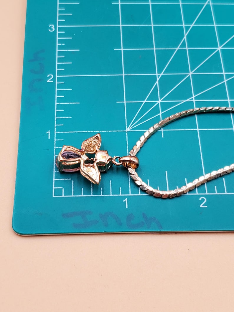 May include: A gold-toned pendant necklace with a floral design. The pendant features a cluster of petals with a central purple gemstone. The chain is a silver-coloured, textured link style. The pendant measures approximately 2.5 cm in length.