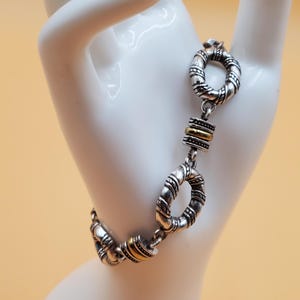 Op de afbeelding: Zilverkleurige armband met een textuurontwerp en goudkleurige accenten. De armband heeft een reeks onderling verbonden cirkelvormige schakels en rechthoekige goudkleurige staven. De sieraden worden getoond op een witte hand tegen een lichtoranje achtergrond.