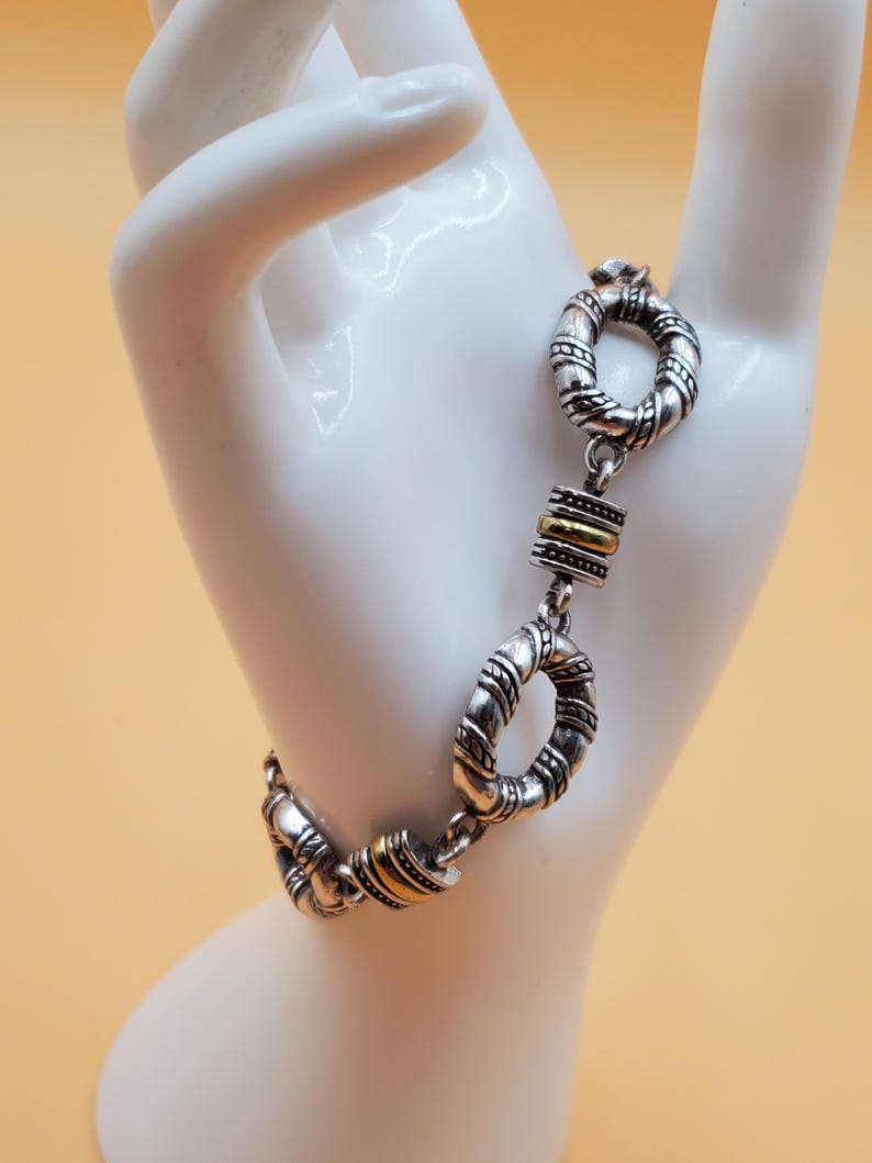 Op de afbeelding: Zilverkleurige armband met een textuurontwerp en goudkleurige accenten. De armband heeft een reeks onderling verbonden cirkelvormige schakels en rechthoekige goudkleurige staven. De sieraden worden getoond op een witte hand tegen een lichtoranje achtergrond.