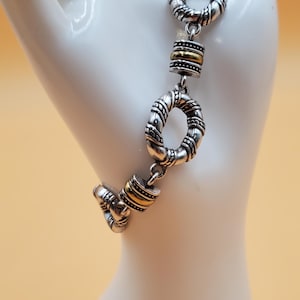 Op de afbeelding: Zilverkleurige armband met gouden accenten. De armband heeft een reeks met elkaar verbonden, getextureerde ringen en cilindrische goudkleurige kralen. Gepresenteerd op een witte mannequinhand tegen een zachtgele achtergrond.