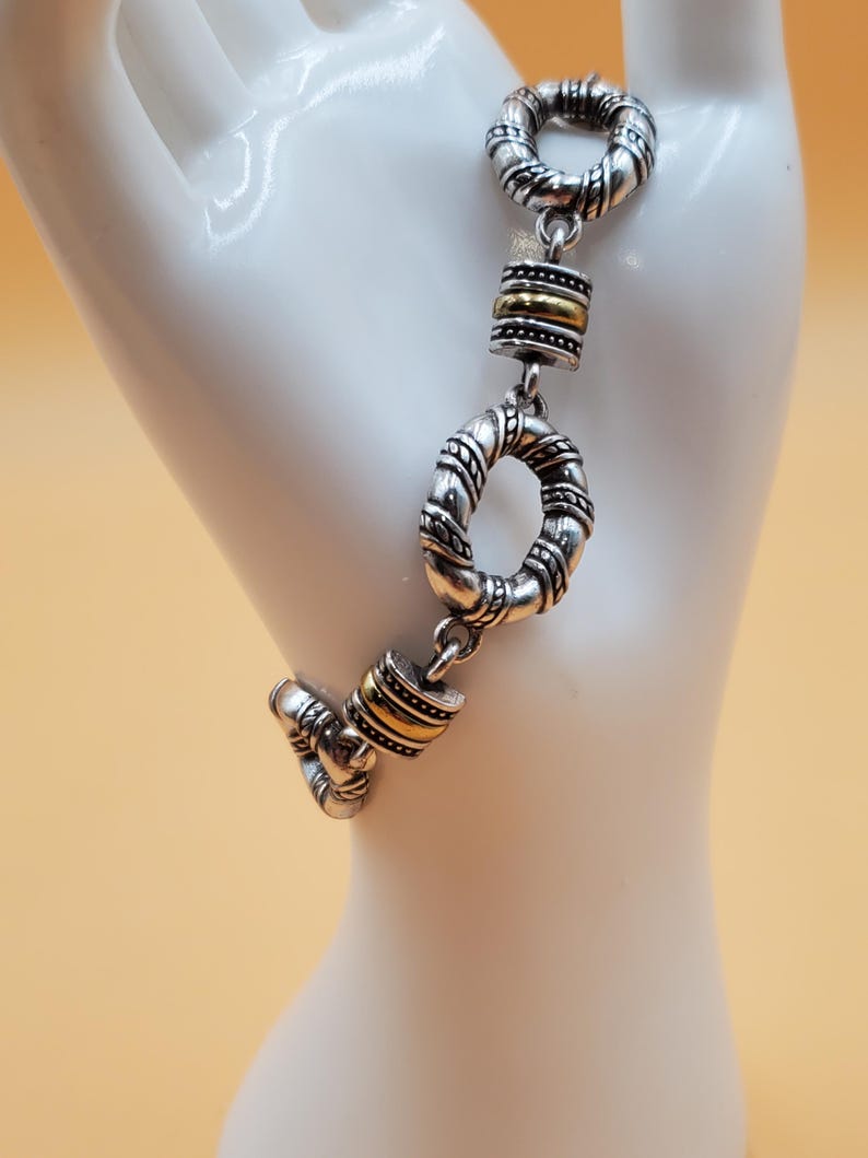 Op de afbeelding: Zilverkleurige armband met gouden accenten. De armband heeft een reeks met elkaar verbonden, getextureerde ringen en cilindrische goudkleurige kralen. Gepresenteerd op een witte mannequinhand tegen een zachtgele achtergrond.