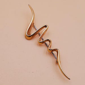 May include: A gold-toned, abstract brooch with a flowing, curved design. The pin has a slender, wavy shape, with a straight pin backing. The brooch is set against a light peach background.