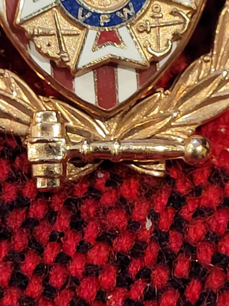 Puede incluir: Un pin de color dorado con un dise&ntilde;o de escudo rojo, blanco y azul. El escudo tiene una estrella en el centro y las letras "U.S.A." sobre la estrella. El escudo est&aacute; rodeado por una corona de hojas doradas y un martillo dorado.