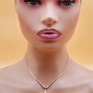 May include: A gold-coloured necklace with an angel pendant. The pendant has a pear-shaped pink gemstone and small clear gemstones. The necklace is shown on a mannequin against a peach-coloured backdrop.