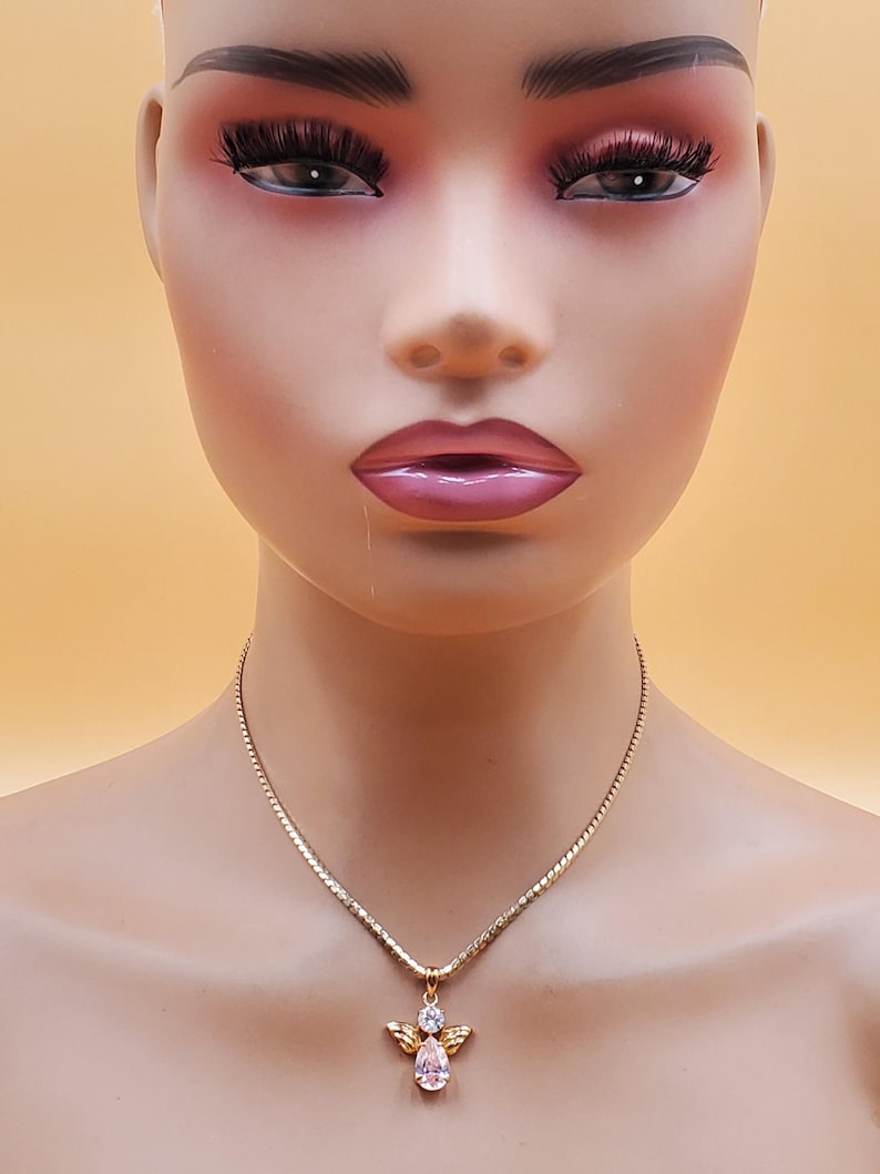 May include: A gold-coloured necklace with an angel pendant. The pendant has a pear-shaped pink gemstone and small clear gemstones. The necklace is shown on a mannequin against a peach-coloured backdrop.