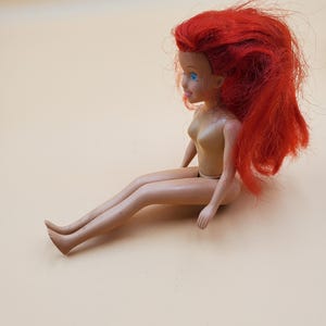 May include: A plastic doll with long, red hair and blue eyes. The doll is sitting with its legs crossed and its arms resting on its lap.