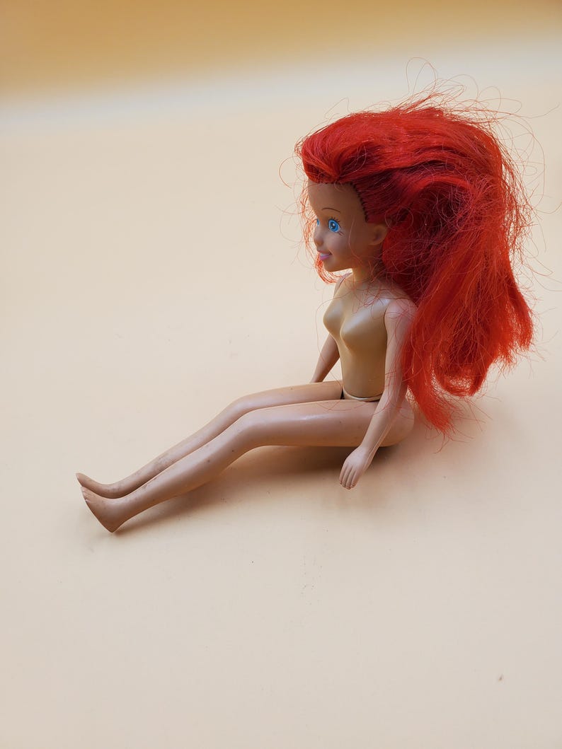 May include: A plastic doll with long, red hair and blue eyes. The doll is sitting with its legs crossed and its arms resting on its lap.