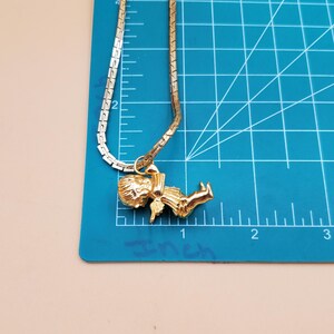Pu&ograve; includere: Una collana color oro con un ciondolo raffigurante una piccola figura. La catena della collana &egrave; a maglie piatte. Il ciondolo &egrave; lungo circa 2,5 cm. La collana &egrave; esposta su un tappetino da taglio blu.