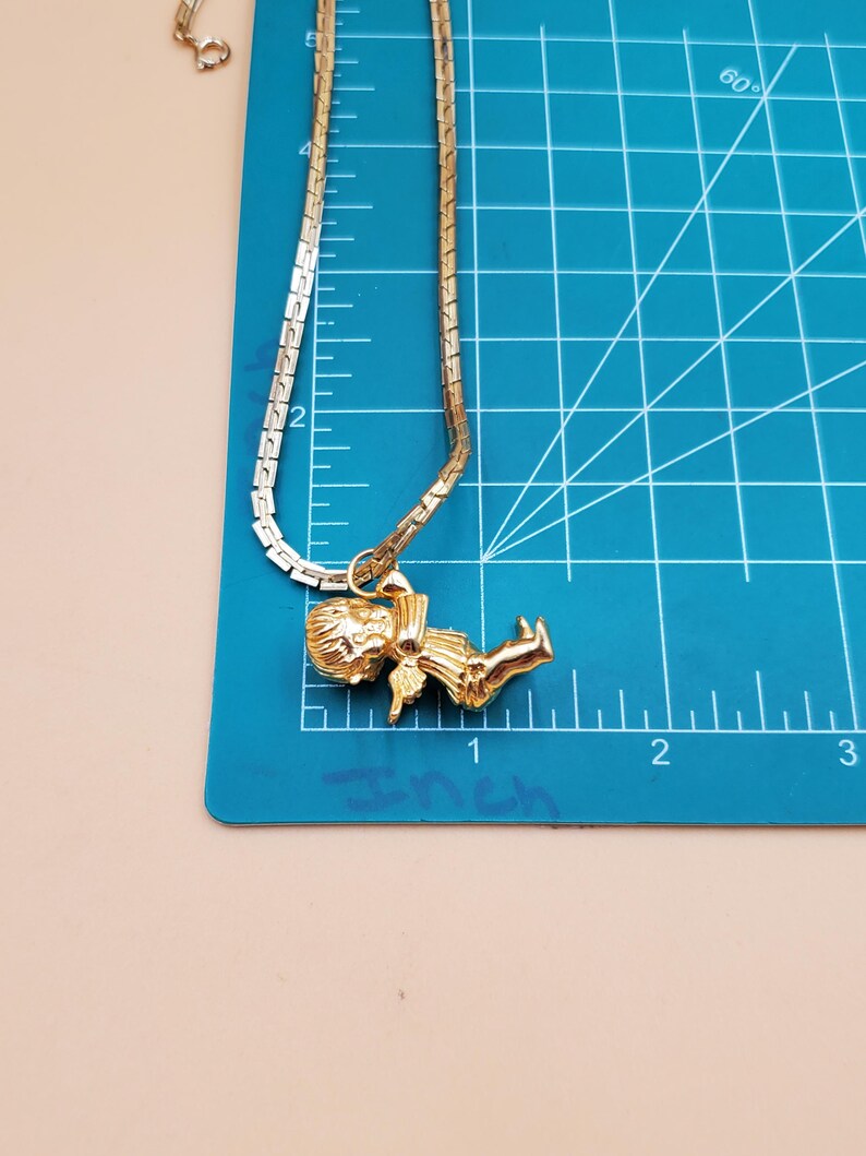 Pu&ograve; includere: Una collana color oro con un ciondolo raffigurante una piccola figura. La catena della collana &egrave; a maglie piatte. Il ciondolo &egrave; lungo circa 2,5 cm. La collana &egrave; esposta su un tappetino da taglio blu.