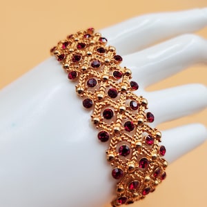 May include: Gold-toned bracelet with a filigree design, adorned with numerous round, deep red gemstones. The bracelet is displayed on a white hand against a soft, peach-colored background. A vintage-inspired piece.