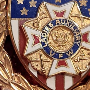 Puede incluir: Pin de tono dorado con un escudo rojo, blanco y azul con una estrella de cinco puntas con un centro rojo, blanco y azul. El escudo est&aacute; rodeado por una corona de hojas doradas. El escudo tiene el texto "LADIES AUXILIARY VFW" en un c&iacute;rculo alrededor de un &aacute;guila calva.