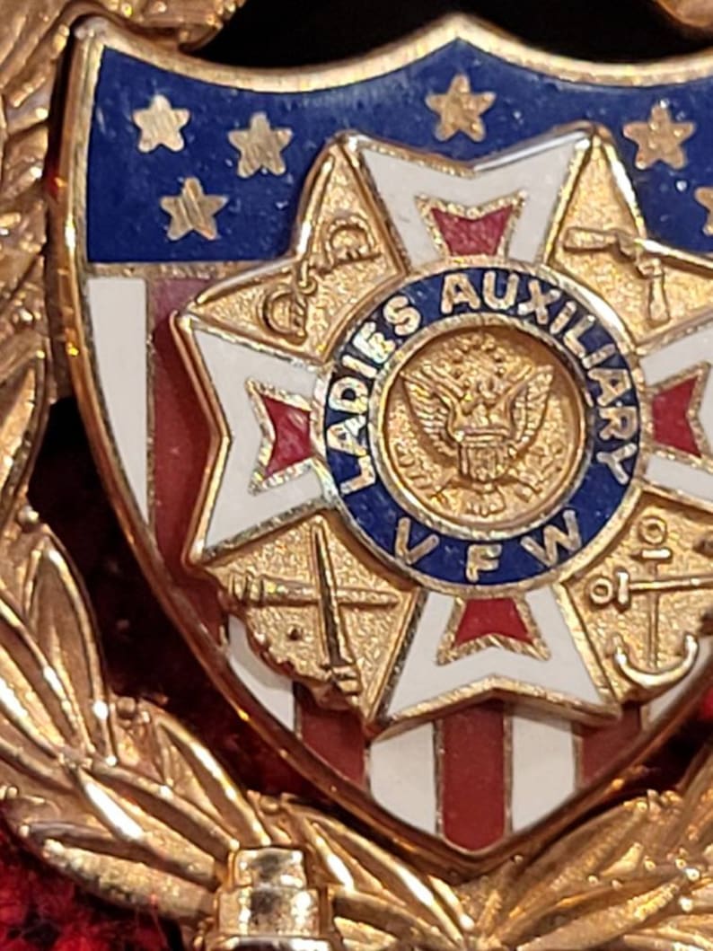 Puede incluir: Pin de tono dorado con un escudo rojo, blanco y azul con una estrella de cinco puntas con un centro rojo, blanco y azul. El escudo est&aacute; rodeado por una corona de hojas doradas. El escudo tiene el texto "LADIES AUXILIARY VFW" en un c&iacute;rculo alrededor de un &aacute;guila calva.
