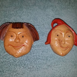 May include: Two wooden carved faces with exaggerated features. The face on the left has brown hair and the face on the right has red hair.