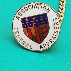 May include: A gold pin with a white circle and the text "Federal Association of Appraisers" around the edge. Inside the circle is a blue shield with three white stars and the letters "A.F.A." in the centre. The shield has red and white stripes.