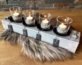 May include: A whitewashed wooden advent candle holder with four glass candle holders and black metal bases. Each holder has a small black slate tag with a number, 1 through 4, written in white chalk. The holder is sitting on a brown faux fur rug.